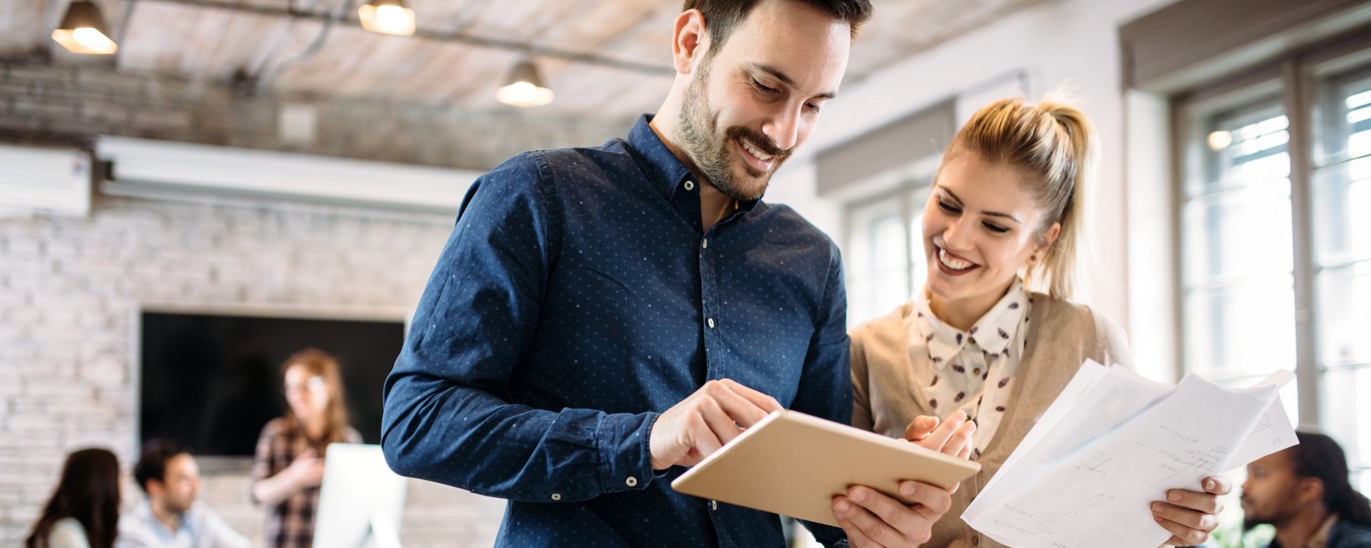 Eine junge Frau und ein junger Mann stehen in einem Bürogebäude und besprechen sich. Beide Personen richten ihren Blick auf eine Tablet, das der dunkelhaarige Mann in seiner Hand hält. Die blonde Dame hält einen Stapel loser Papiere in der Hand und zeigt lächelnd auf das Tablet. Im Hintergrund ist eine Büroumgebung mit weiteren Personengruppen zu sehen, die alle miteinander kommunizieren.