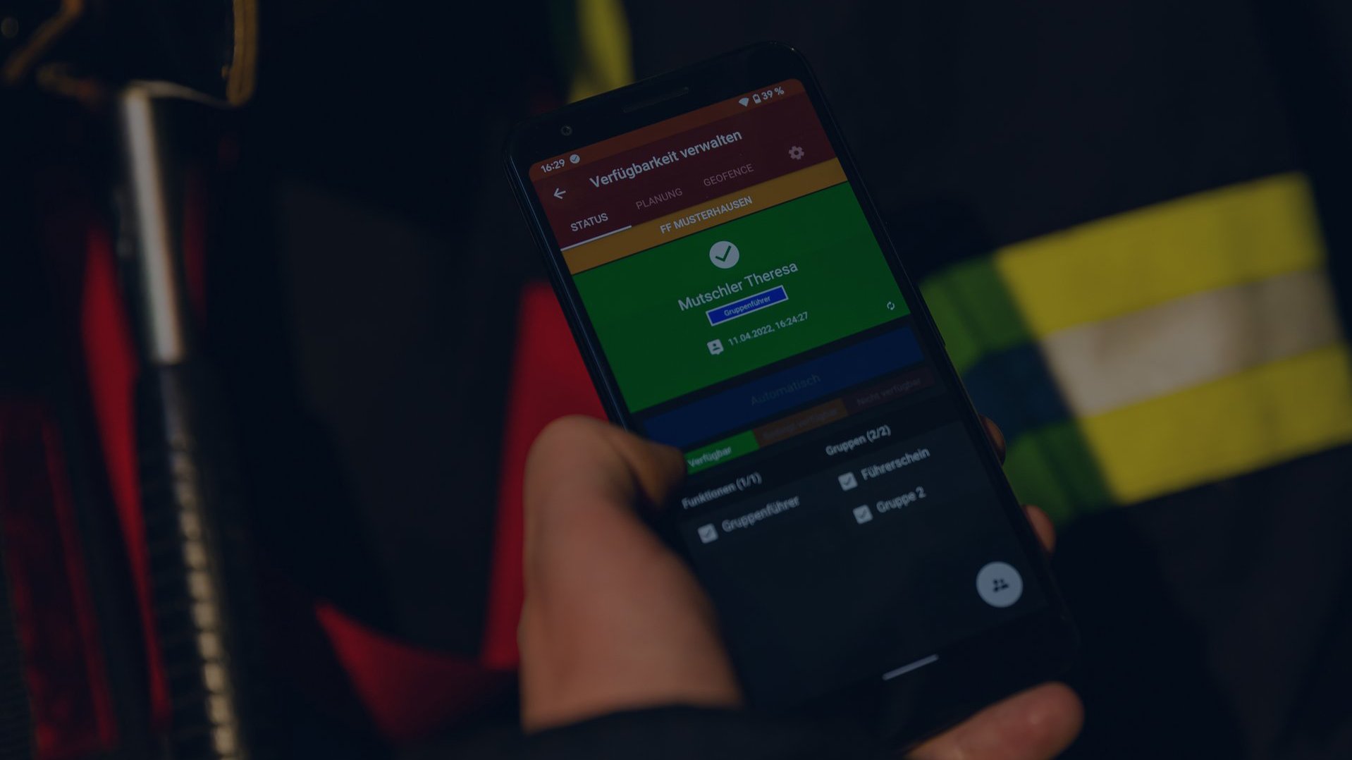 Nahaufnahme eines Smartphones, welches von einer Einsatzkraft gehalten wird und die Verfügbarkeitsverwaltung der Alamos Software-Lösungen zeigt. Im Hintergrund sind unscharf Einsatzklamotten mit gelben Leuchtstreifen zu erkennen. Das Bild wurde mit einer vollflächigen dunkelblauen Überlagerung abgedunkelt.