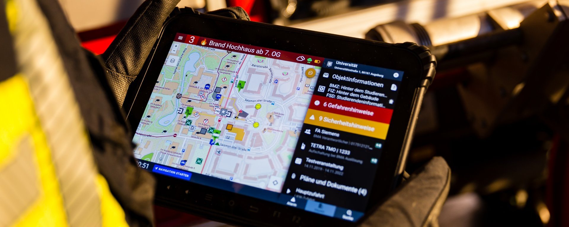 Nahaufnahme eines Einsatztablets, das von einer Einsatzkraft in Schutzkleidung gehalten wird und die Kartenübersicht für Gefahrenhinweise der Alamos Software-Lösungen zeigt. Im Hintergrund ist unscharf die Seite eines Feuerwehrfahrzeugs zu sehen.