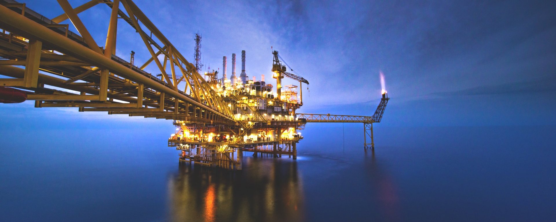 Darstellung einer Industrieplattform auf dem Wasser. Die Perspektive ist vom Land aus wodurch auf dem Bild nur die Industrieplattform umgeben von Wasser zu sehen ist. Die Aufnahme enstandt bei Nacht und die Plattform ist mit gelben und roten Lichtern beleuchtet.