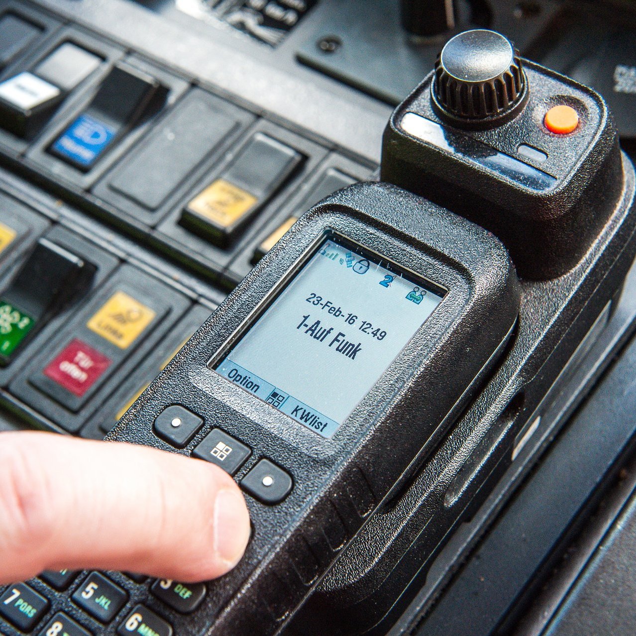 Nahaufnahme eines Funkgeräts innerhalb eines Einsatzfahrzeugs. Eine Person tippt mit ihrem Finger auf einen der Knöpfe des Geräts während der Display einen Anruf auf Funk anzeigt. Im Hintergrund ist ein Board mit vielen Schaltern und Hebeln zu sehen, von denen einige aktiviert sind.