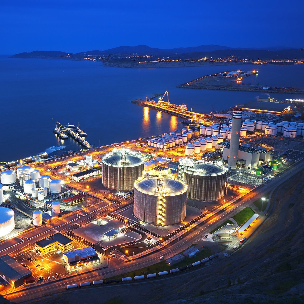 Schräge Vogelperspektive einer großen Industrieplattform umgeben von Gewässer bei Nacht. Die sichtbaren Gebäude und Türme sind mit gelben und roten Lichtern und Lampen beleuchtet.