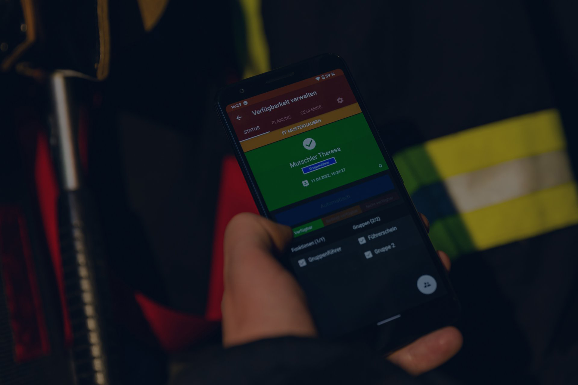 Nahaufnahme eines Smartphones, welches von einer Einsatzkraft gehalten wird und die Verfügbarkeitsverwaltung der Alamos Software-Lösungen zeigt. Im Hintergrund sind unscharf Einsatzklamotten mit gelben Leuchtstreifen zu erkennen. Das Bild wurde mit einer vollflächigen dunkelblauen Überlagerung abgedunkelt.